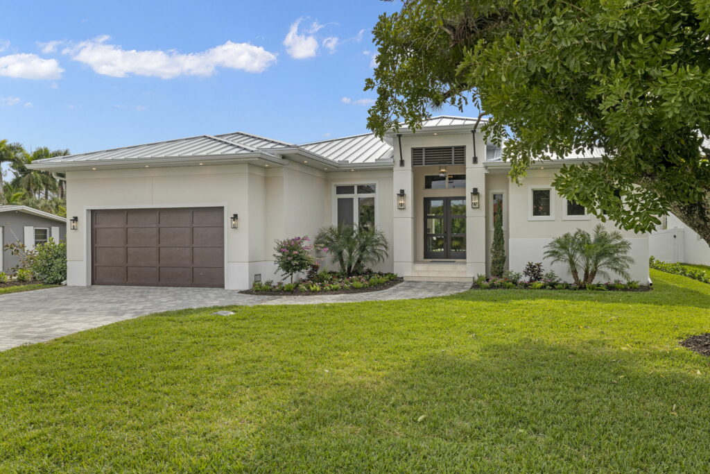 View of a white single-story house with a brown garage door, white walls, a square paver driveway, and manicured yard from the front right of the house